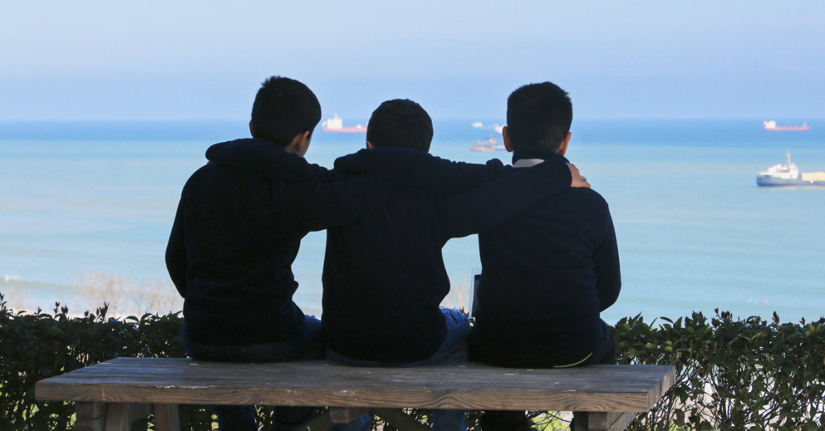 3 boys from behind sitting side by side face the ocean